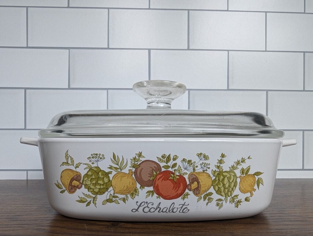 A vintage CorningWare white ceramic baking dish with a glass lid sits on a wooden surface. The dish features illustrations of assorted fruits and vegetables and the word "L'Echalote" on its side. The background is adorned with white tiles.