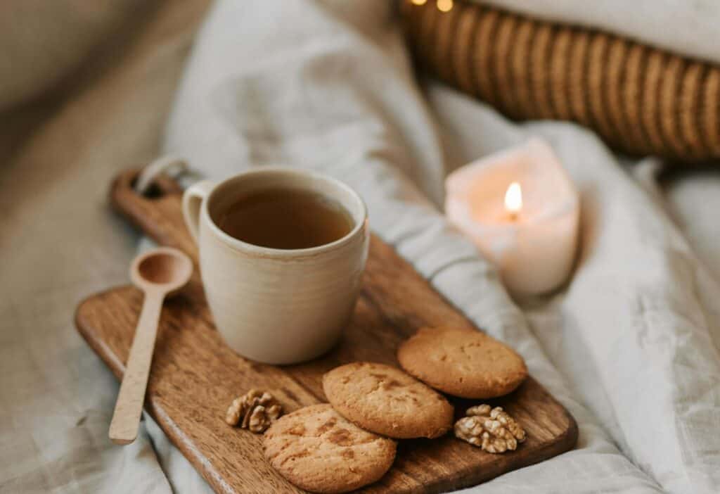 A cozy living setting with a cup of tea, cookies, and walnuts on a wooden tray next to a lit candle, all arranged on a bed with soft linen.