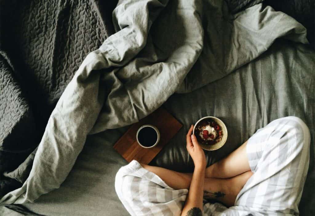 A person sitting on a bed holding a bowl of yogurt with berries, with a book and a cozy grey blanket nearby.