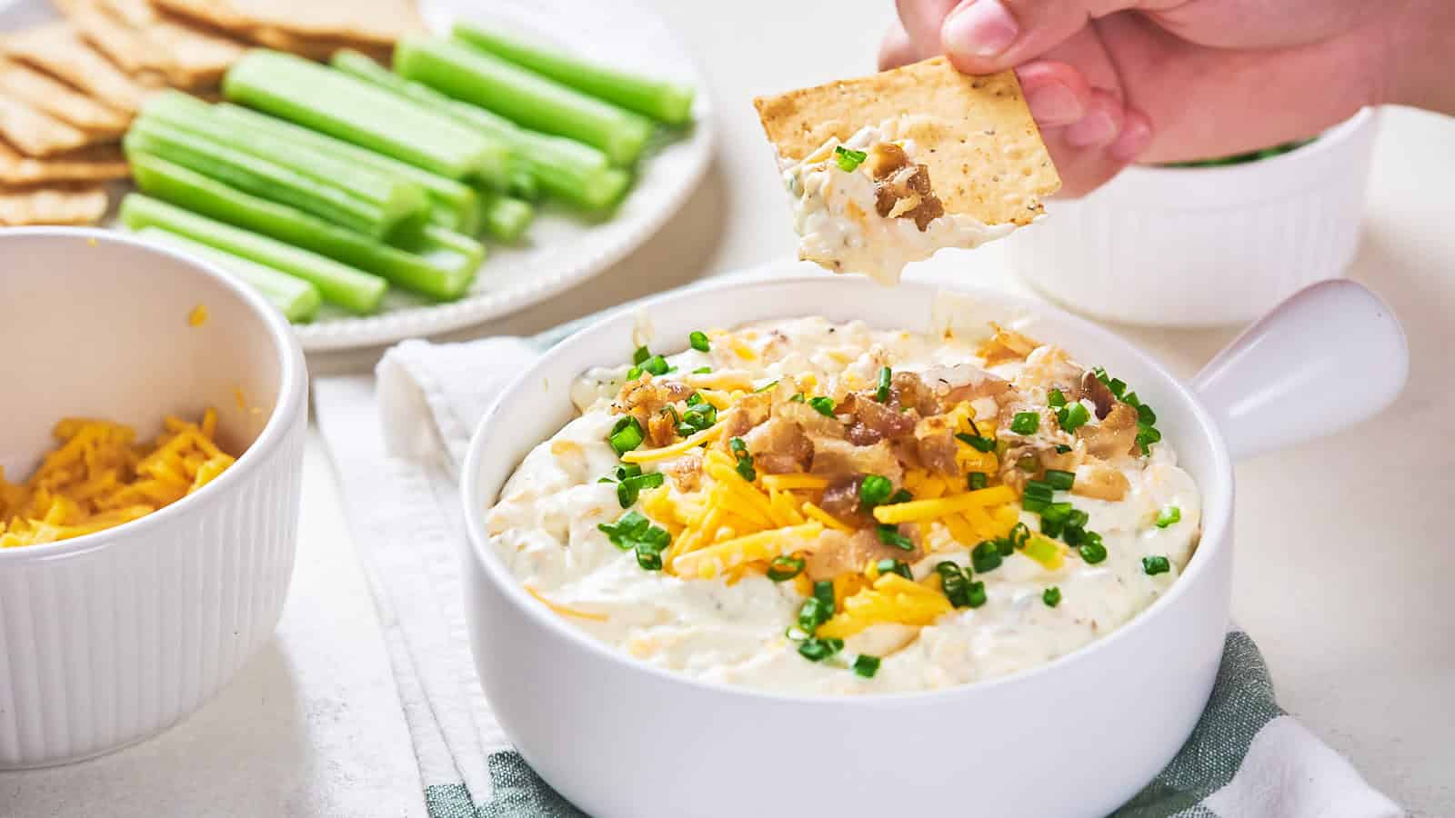 A person dipping a cracker into a bowl of creamy dip topped with chives and shredded cheese, with celery sticks and additional crackers on the side.