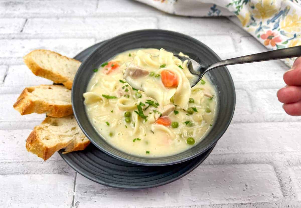 A hand holding a spoonful of creamy chicken noodle soup with vegetables over a bowl, accompanied by slices of bread on a plate.