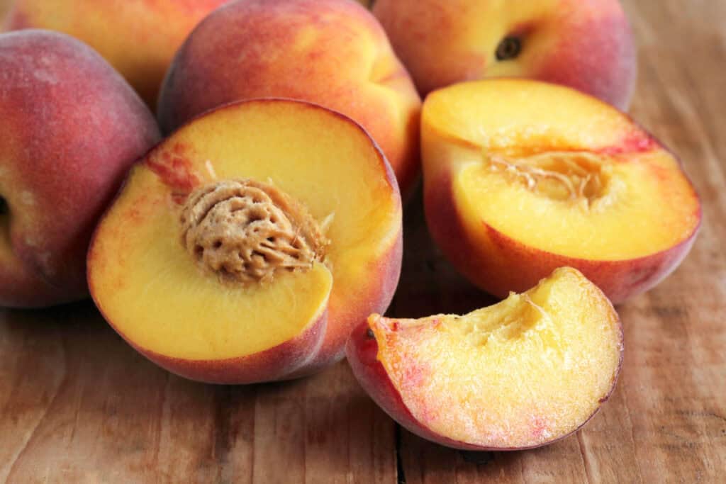 A group of ripe peaches on a wooden surface, with one peach sliced open to reveal the pit and a wedge cut out.