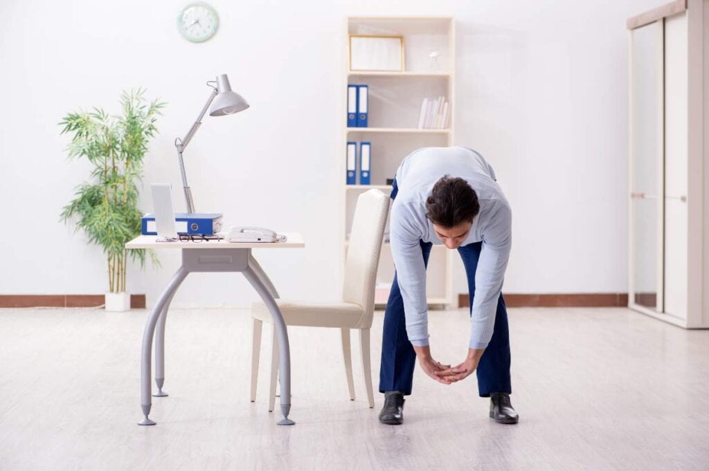 A person in business attire is bending forward to touch their toes in an office setting, next to a desk and chair. The office is furnished with a lamp, plant, shelves, and a clock on the wall.