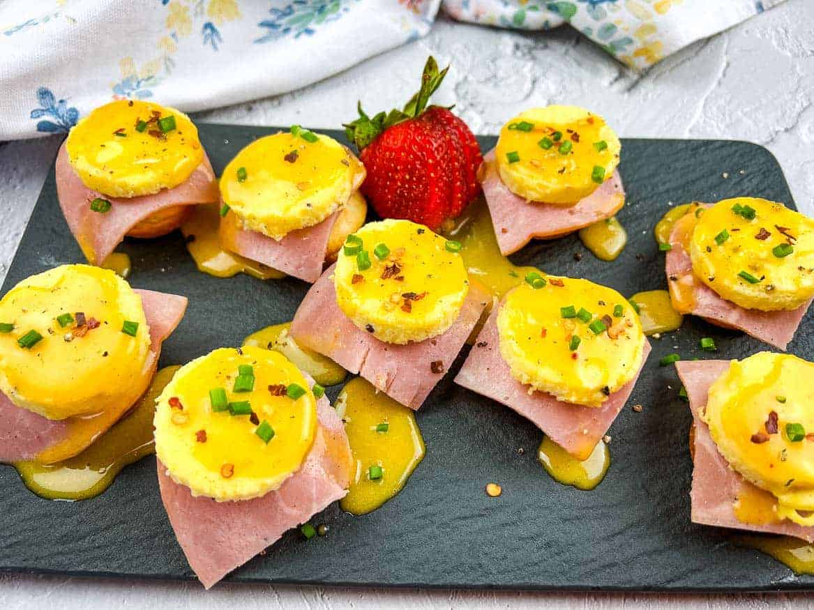 A slate platter with eggs benedict bites garnished with chopped chives and a strawberry in the center.
