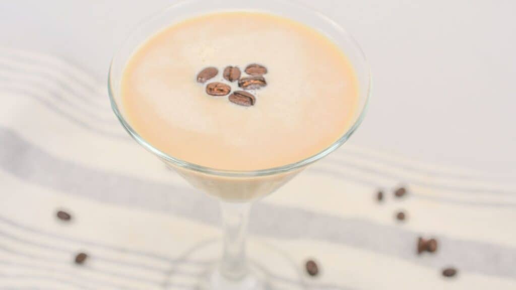 A creamy coffee drink in a martini glass, topped with coffee beans, on a striped cloth with scattered beans around.