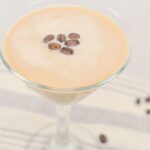 A creamy coffee drink in a martini glass, topped with coffee beans, on a striped cloth with scattered beans around.