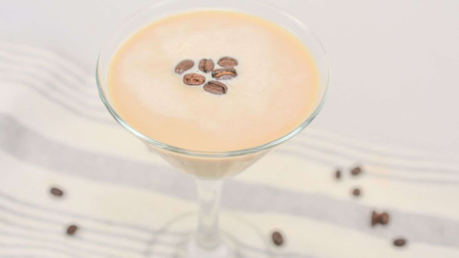 A creamy coffee drink in a martini glass, topped with coffee beans, on a striped cloth with scattered beans around.