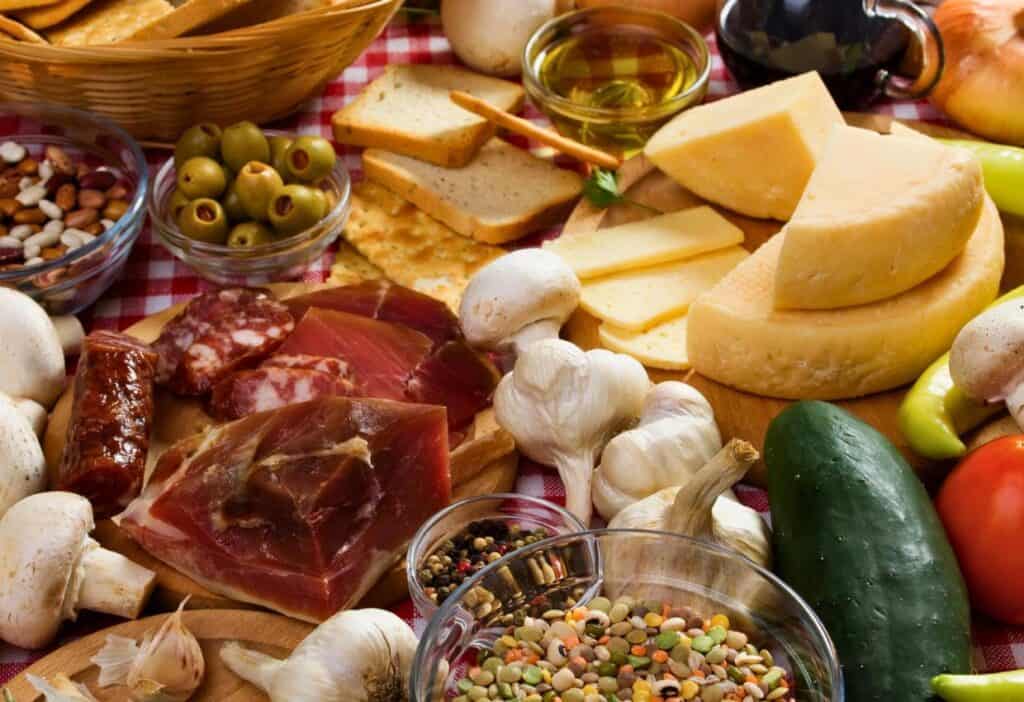 A variety of European foods, including cheese, bread, ham, olives, mushrooms, garlic, cucumber, tomato, nuts, and grains, displayed on a red-checked tablecloth.