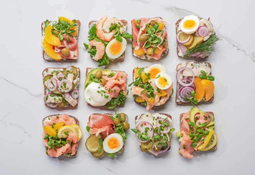 A neatly arranged assortment of open-faced sandwiches topped with various ingredients, including eggs, shrimp, smoked salmon, vegetables, and greens, on a white marble surface showcases a delightful array of European food.