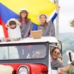 A happy family of four, dressed in striped tops, enjoying a picnic on top of a red vintage car under the colombian flag in a scenic landscape.