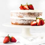 A naked cake with white frosting between layers, garnished with fresh strawberries on top and around the base, displayed on a white stand with a light background.