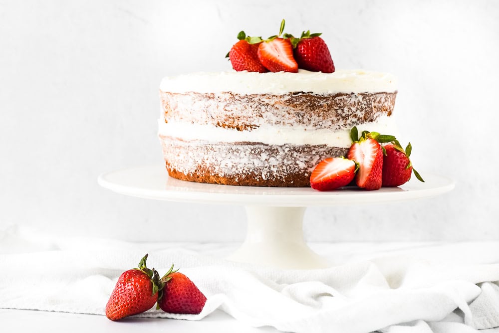 A naked cake with white frosting between layers, garnished with fresh strawberries on top and around the base, displayed on a white stand with a light background.