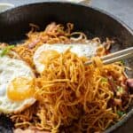 A bowl of fried noodles topped with two sunny-side-up eggs. A pair of chopsticks is lifting a portion of the noodles.