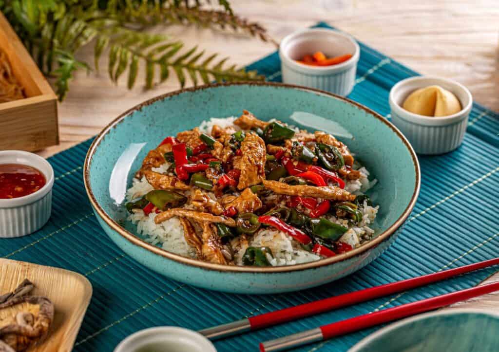 A vibrant dish of rotisserie chicken with bell peppers and green beans served over rice in a turquoise bowl, accompanied by red chopsticks and various side dishes.