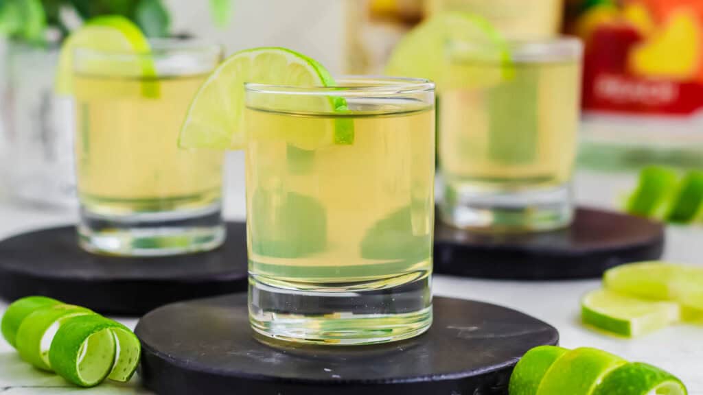 Three shots of golden tequila served in clear shot glasses, each garnished with a lime wedge, on black coasters with lime peels scattered around.