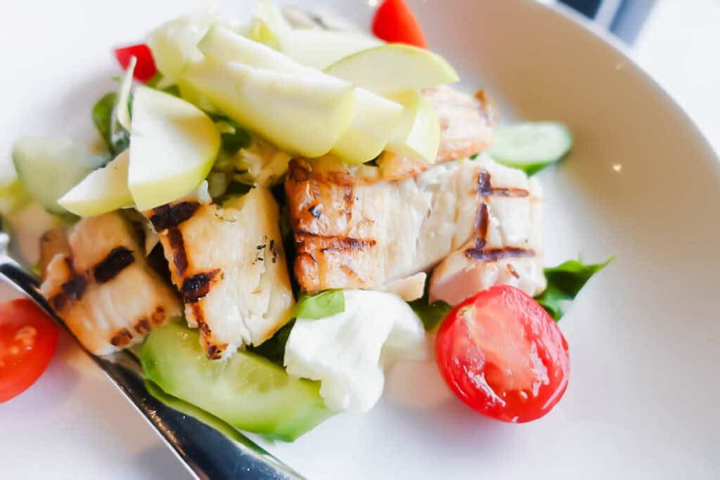 A salad with grilled chicken, sliced green apples, cherry tomatoes, cucumber slices, and a dollop of creamy dressing in a white bowl. A fork is placed in the bowl.