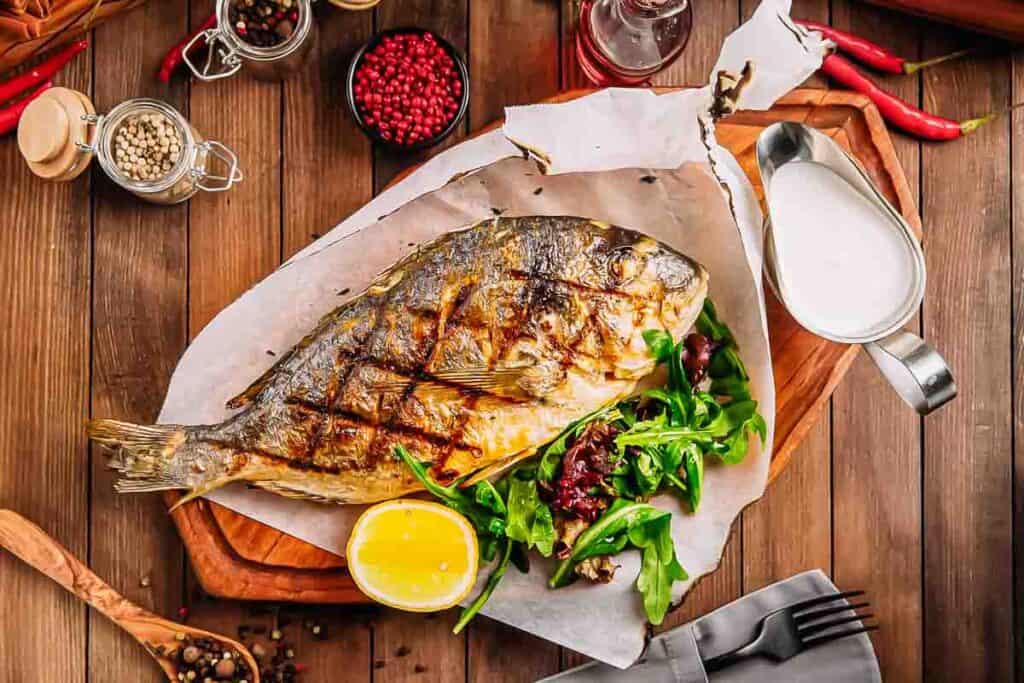 Grilled fish served on a wooden board with greens, half a lemon, and a sauce jug on the side. Surrounding items include peppercorns, spices, and utensils on a wooden table.