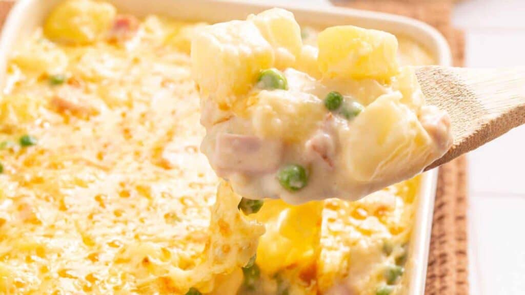 A creamy potato casserole with peas, topped with melted cheese, being served with a wooden spoon.
