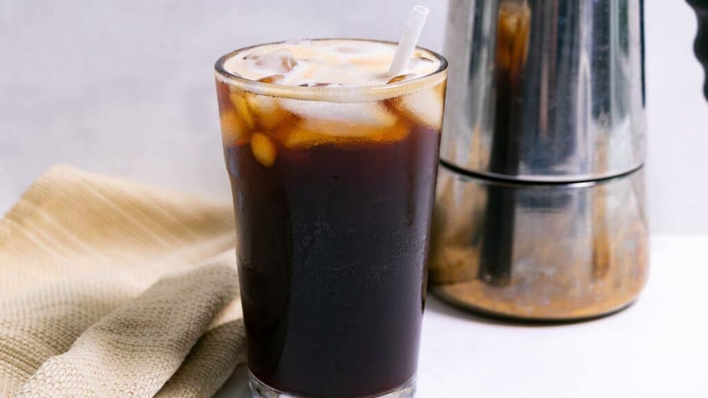 A glass of iced coffee with a straw, next to a coffee maker on a kitchen counter with a beige napkin.