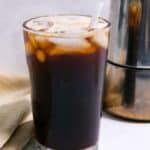 A glass of iced coffee with a straw, next to a coffee maker on a kitchen counter with a beige napkin.