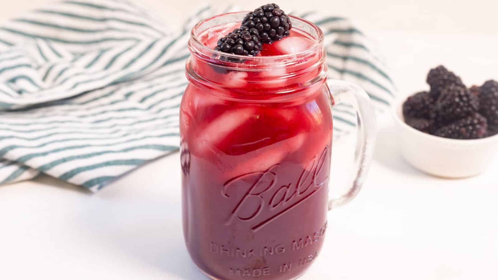 A glass mason jar filled with a red berry drink and topped with blackberries, accompanied by a small bowl of blackberries and a striped napkin on a white surface.