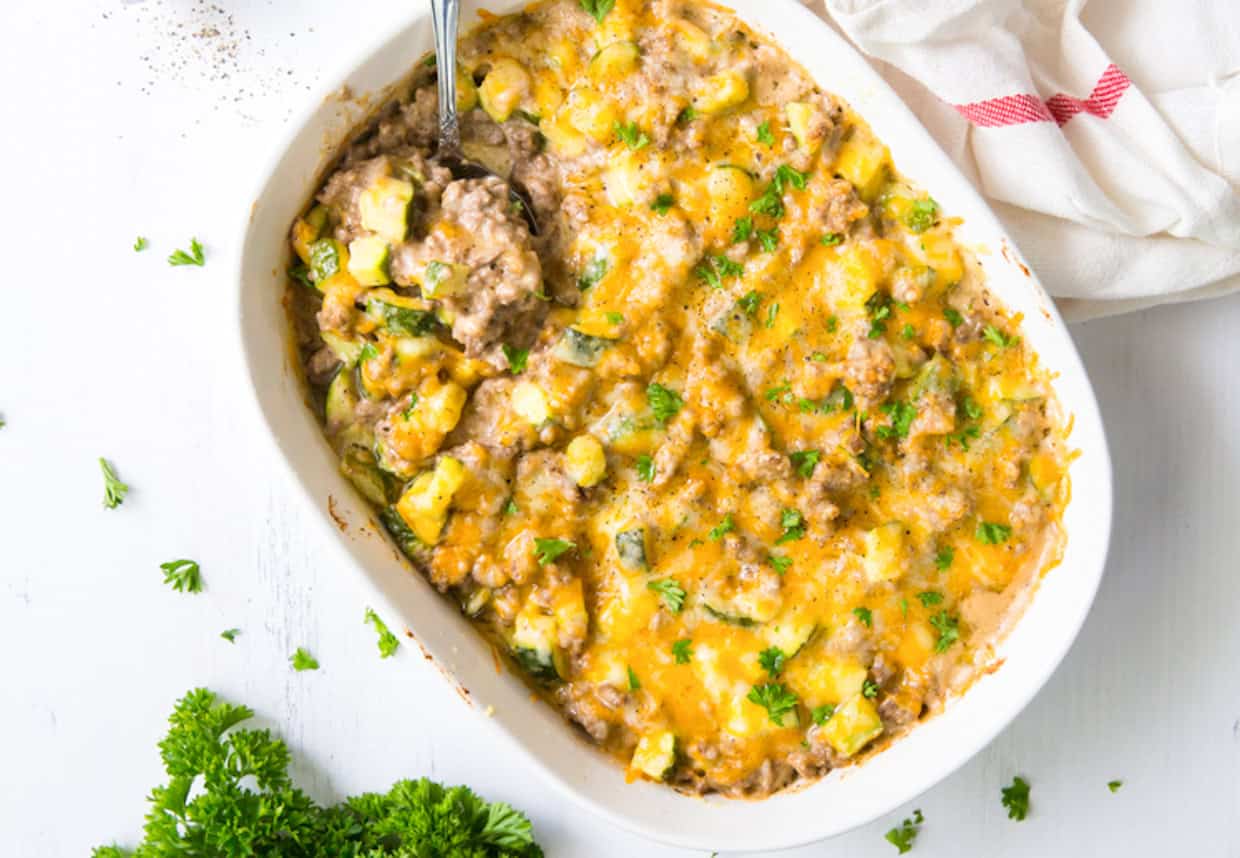 A casserole dish containing a ground meat and vegetable mixture topped with melted cheese and garnished with herbs. A spoon is scooping out a portion. Fresh parsley is nearby.