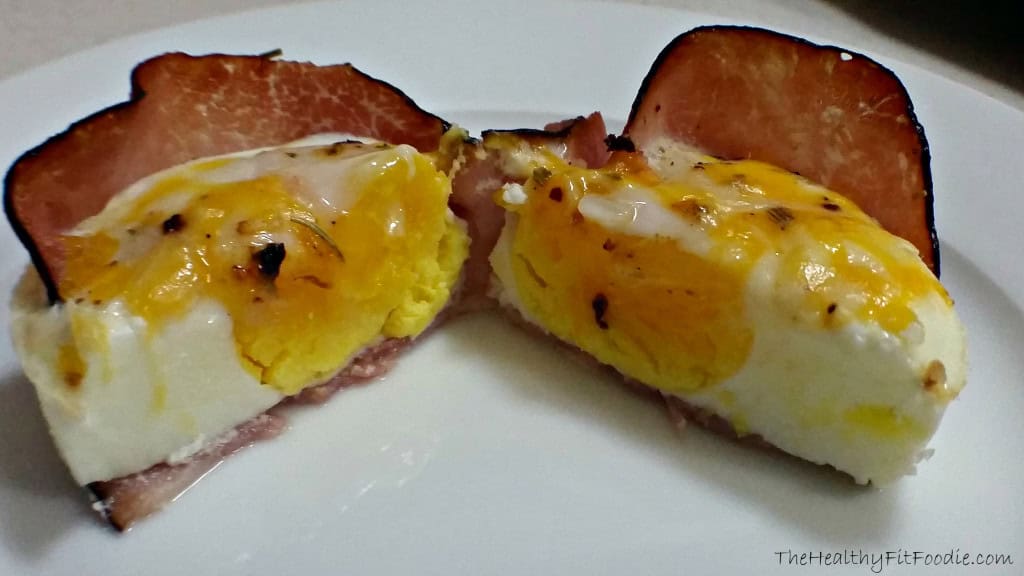 Two breakfast cups made from crispy ham filled with soft-cooked eggs and melted cheese on a white plate.