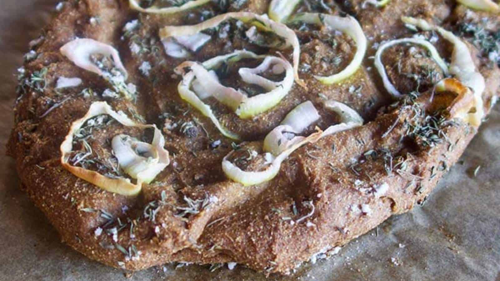 Close-up of a homemade focaccia bread with onions and herbs on a textured surface.