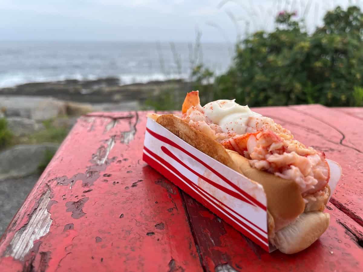 A Maine lobster roll in a red and white striped container sits on a weathered red picnic table with a coastal scene visible in the background.
