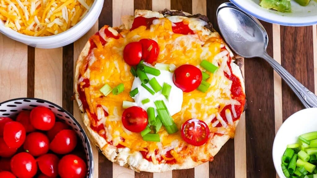 Homemade mini pizza topped with cheese, tomatoes, green onions, and a dollop of sour cream, surrounded by bowls of ingredients.