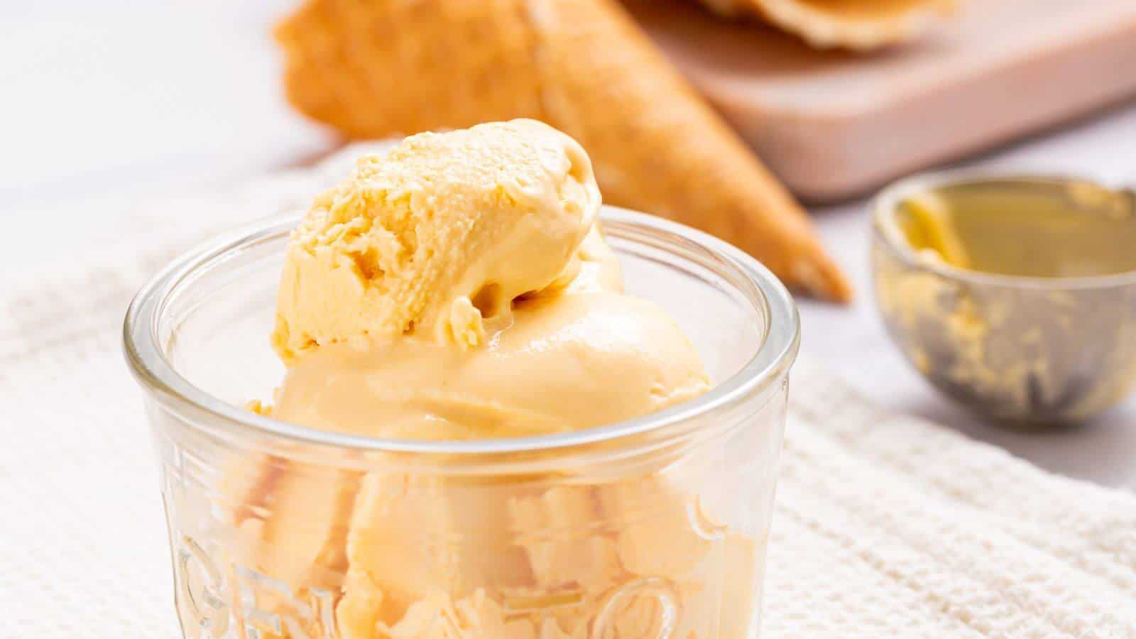A scoop of no-churn vanilla ice cream in a glass jar, with a waffle cone and a wooden spoon in the background on a white tablecloth.