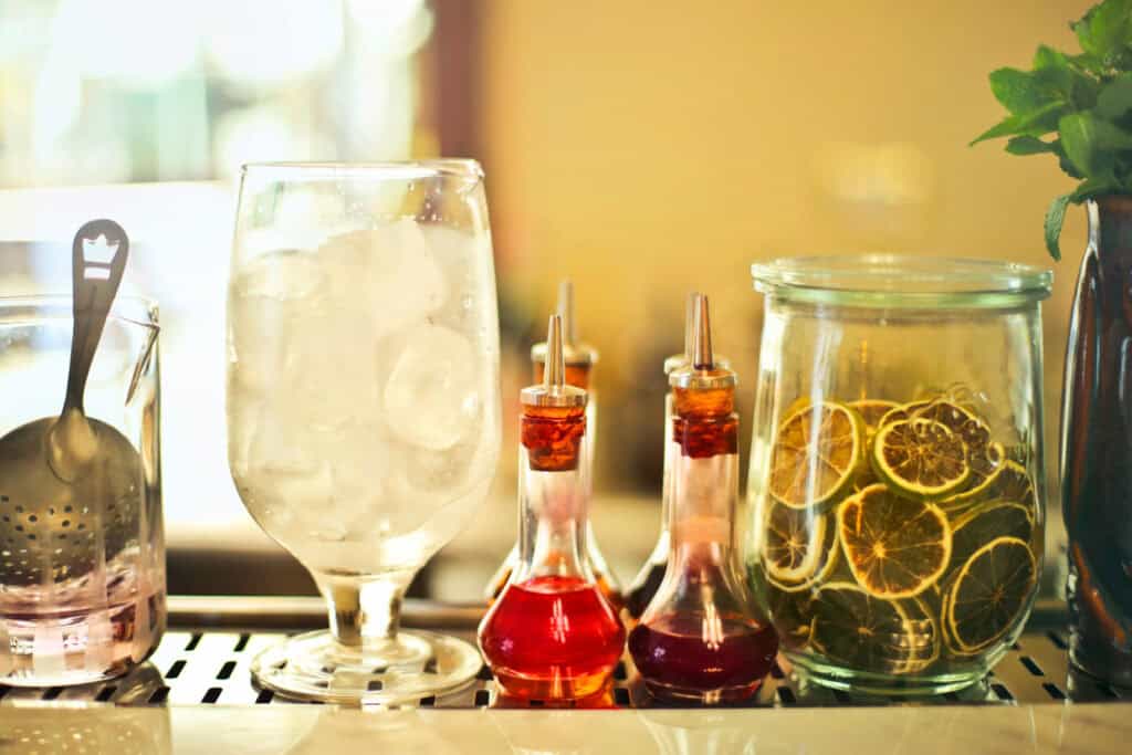 A glass of ice next to a jar of citrus slices and bottles of colorful syrups on a bar counter.