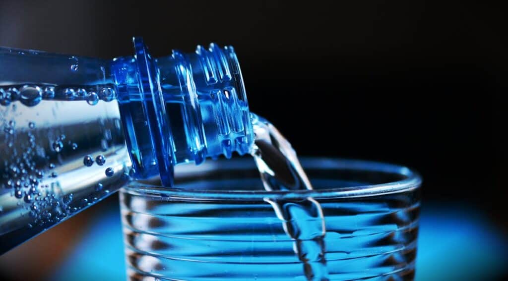 A blue plastic bottle is pouring water into a clear, rippled glass.