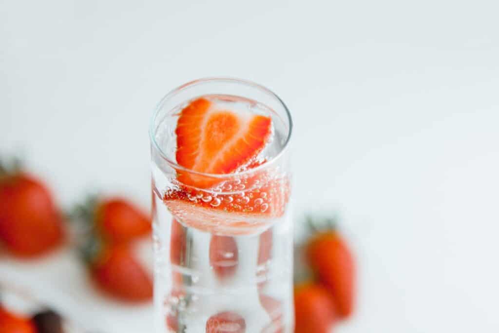 A glass of sparkling water with a sliced strawberry floating inside, surrounded by whole strawberries in the background.