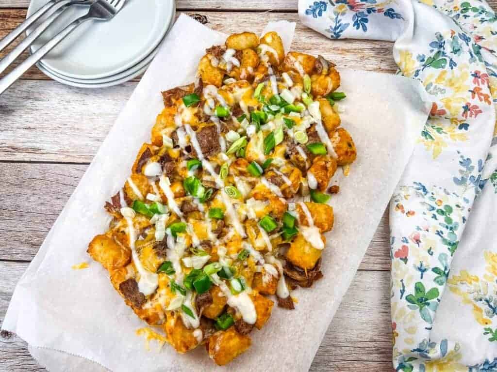 A tray of loaded tater tots topped with Alfredo sauce, scallions, and green onions on a wooden table next to plates and utensils.