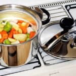 A stainless steel pot with mixed vegetables on a gas stove, next to another pot with a closed lid.