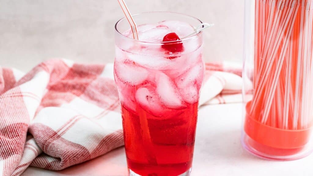 A glass of bright red cherry soda with ice cubes and a straw, accompanied by a red and white striped straw pack and a checkered napkin on a light background.