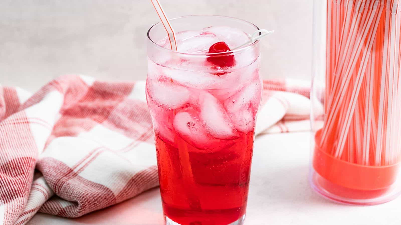 A glass of red soda with ice and a cherry, accompanied by a straw and a red and white napkin, on a light background.