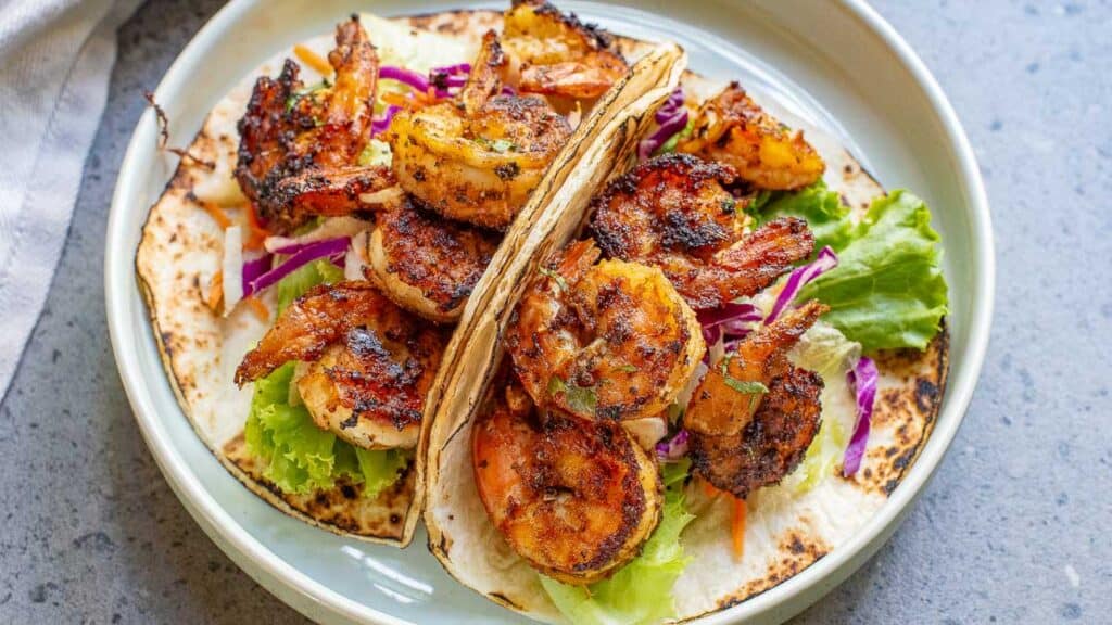 Grilled shrimp tacos with purple cabbage and lettuce on a white plate, served over tortillas.