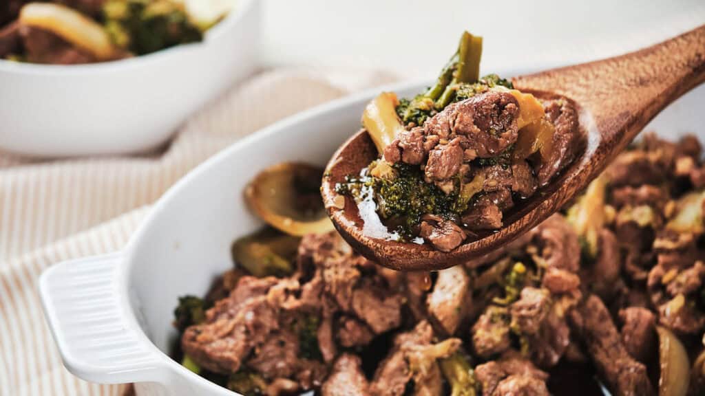 A wooden spoon scoops beef and broccoli from a white casserole dish, showcasing the meal's rich sauce and tender chunks of meat.