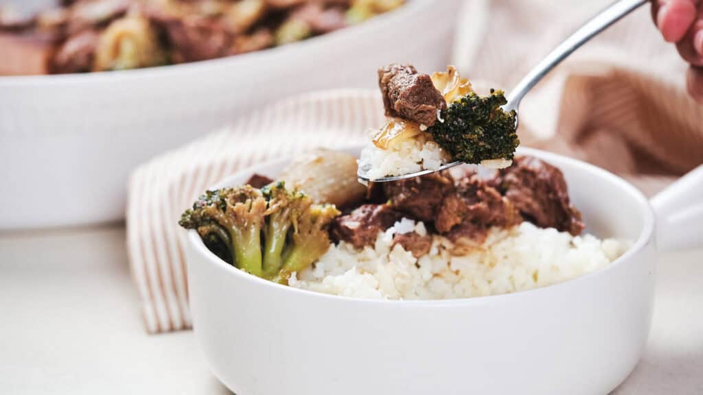 A person's hand serving broccoli and beef over a bowl of rice with a fork.