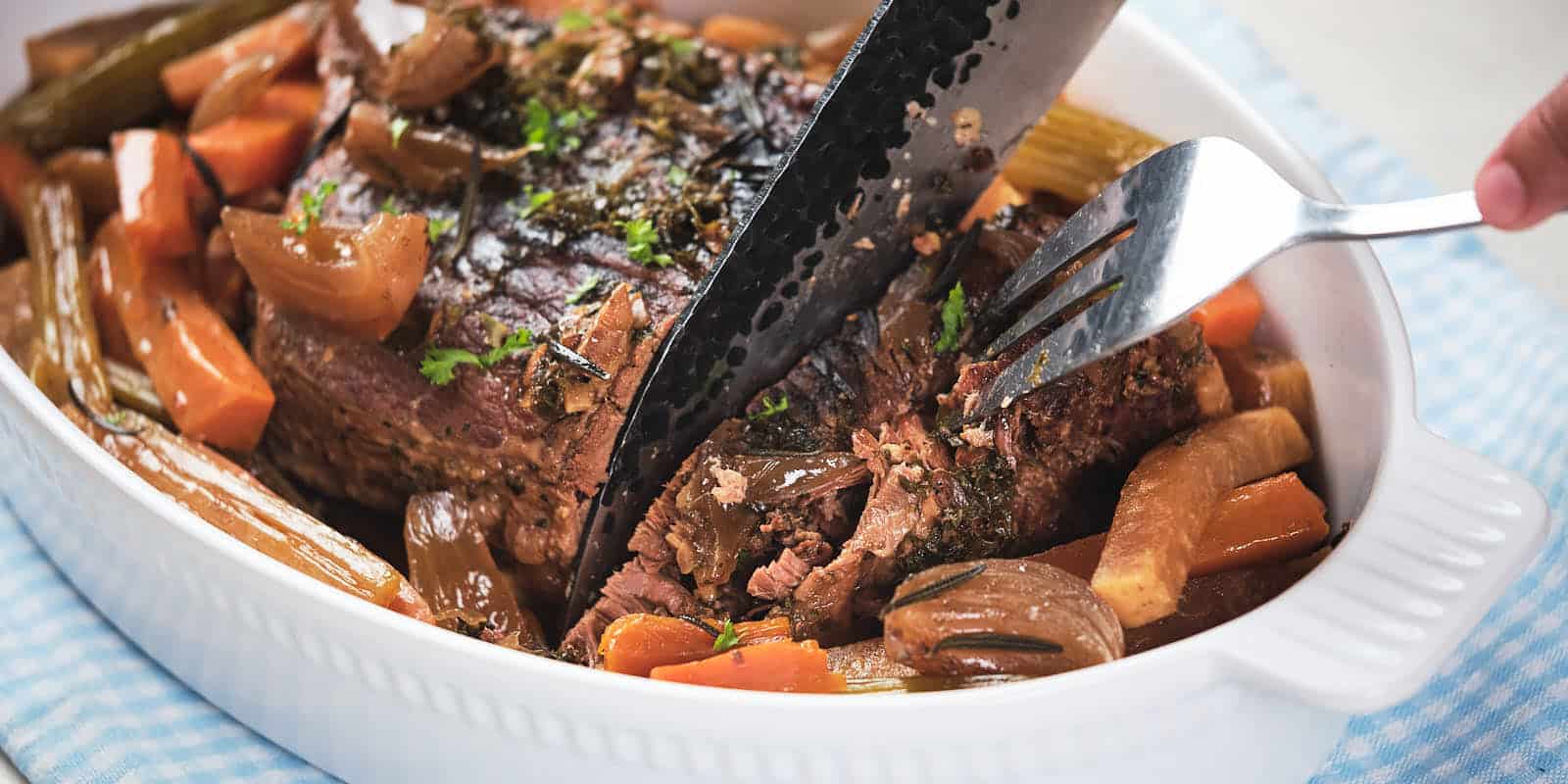 A person slicing a pot roast with carrots and onions in a white serving dish, using a carving fork and knife.