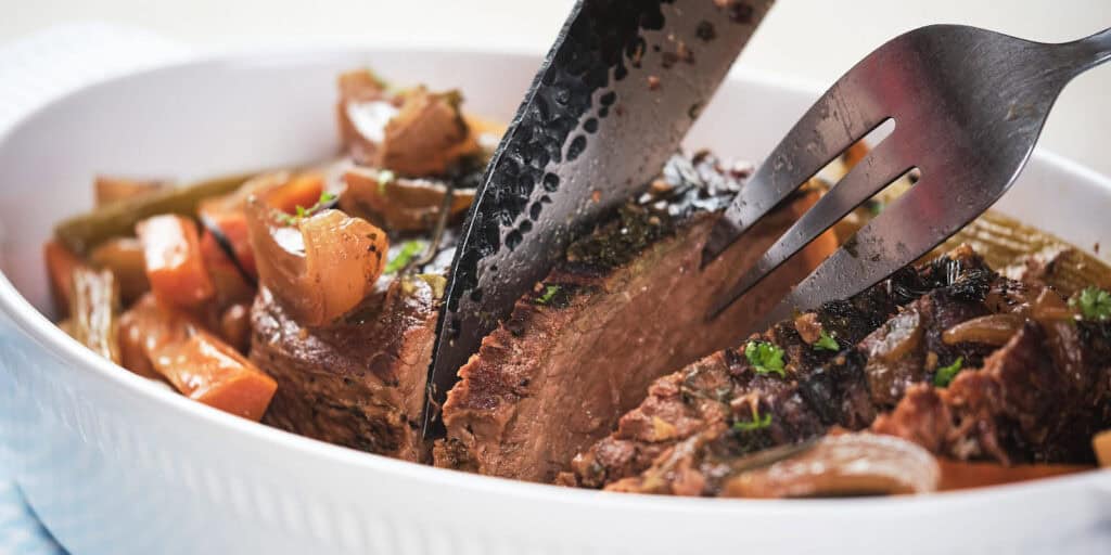 Carving fork and knife slicing into braised beef brisket in a white serving dish with carrots and onions.