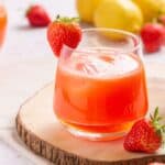 A fresh strawberry lemonade served on a wooden coaster, garnished with a whole strawberry, with lemons and strawberries in the background.