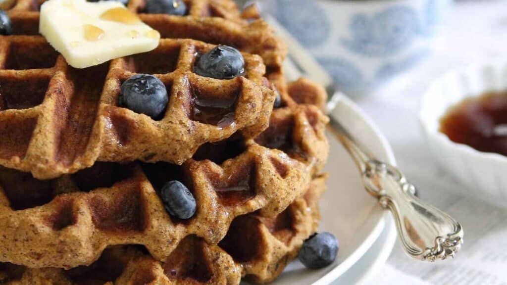 A stack of waffles topped with blueberries, a pat of butter, and syrup on a white plate with a silver fork next to a cup and a bowl of extra syrup.