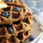 A stack of waffles topped with blueberries, a pat of butter, and syrup on a white plate with a silver fork next to a cup and a bowl of extra syrup.