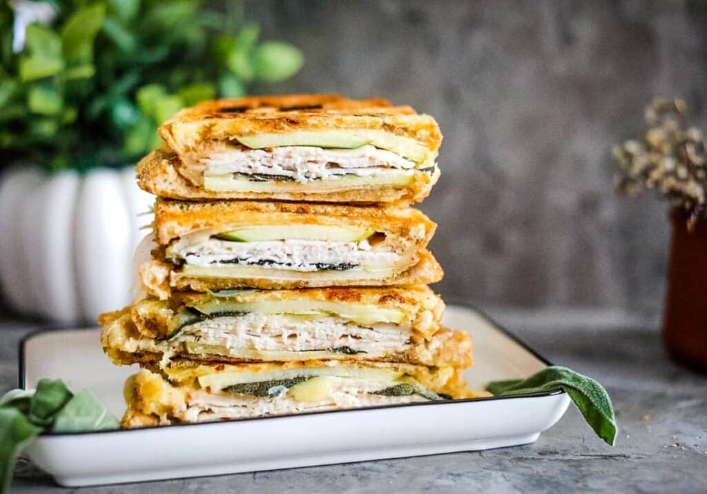 A stack of grilled sandwiches with layers of turkey, cheese, and pickles, served on a white plate with a blurred background of green plants and a brown vase.