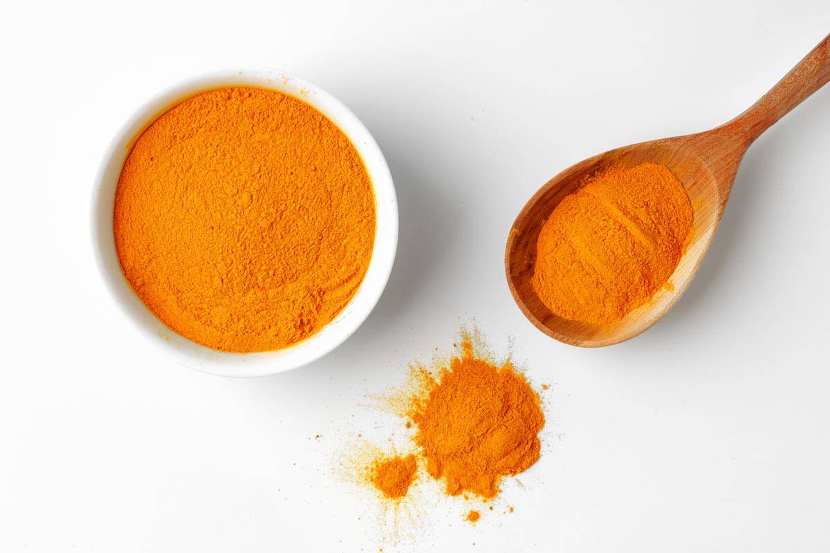 A bowl filled with turmeric powder next to a wooden spoon with turmeric, and some spilled on a white surface.