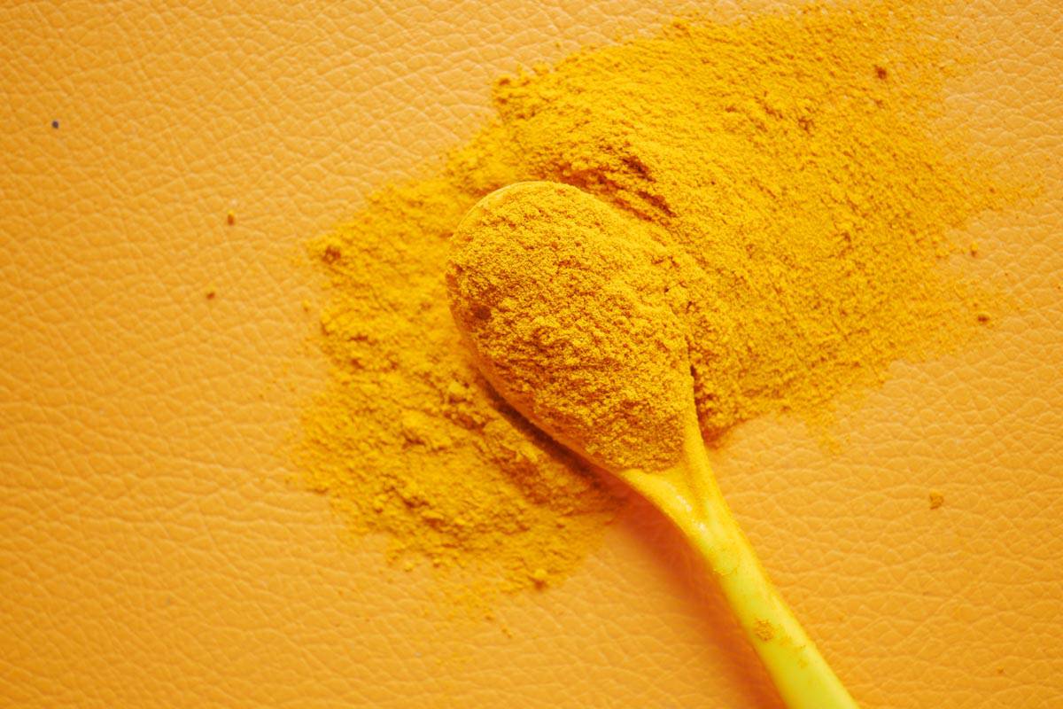 A wooden spoon covered in yellow turmeric powder on an orange textured surface.