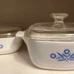 Two vintage CorningWare white ceramic baking dishes with clear glass lids are placed on a shelf. The dishes have blue flower designs on the sides.